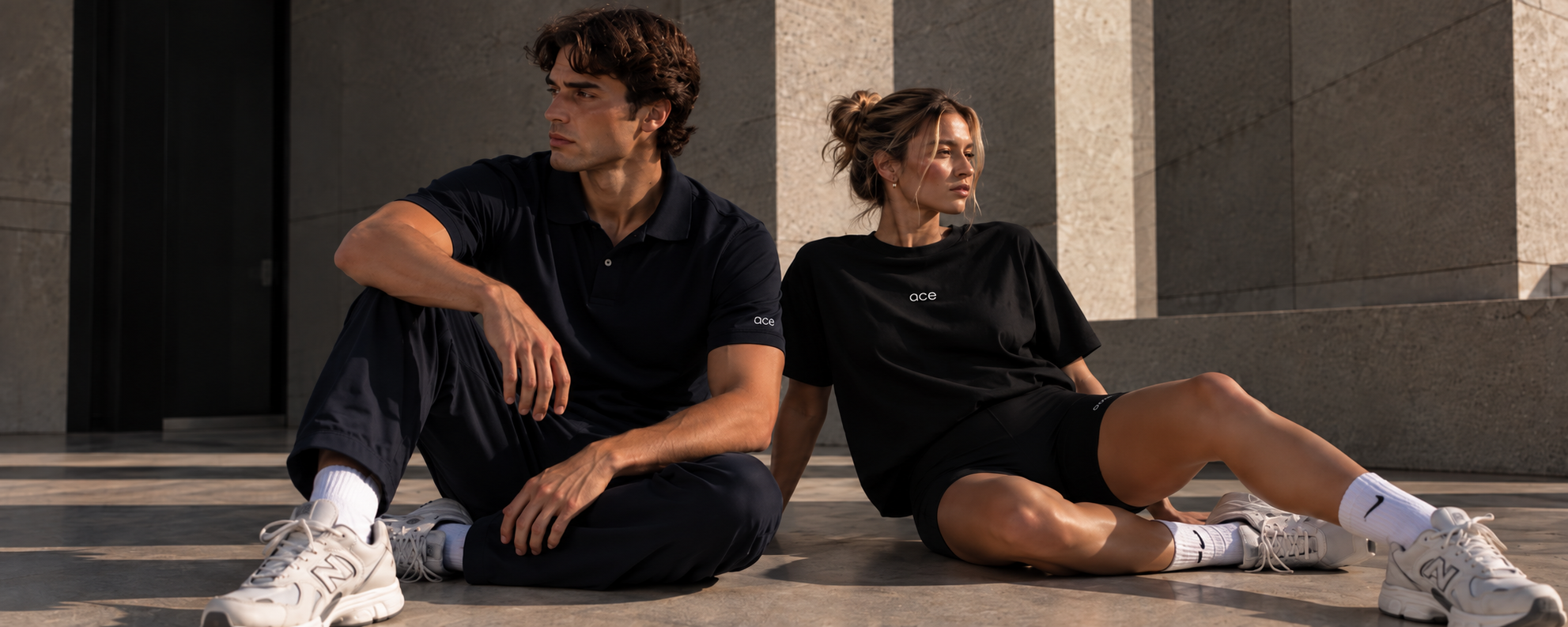 Two models seated in sunlit stone architecture wearing a navy Ace Club polo and a black Ace Club t-shirt with shorts